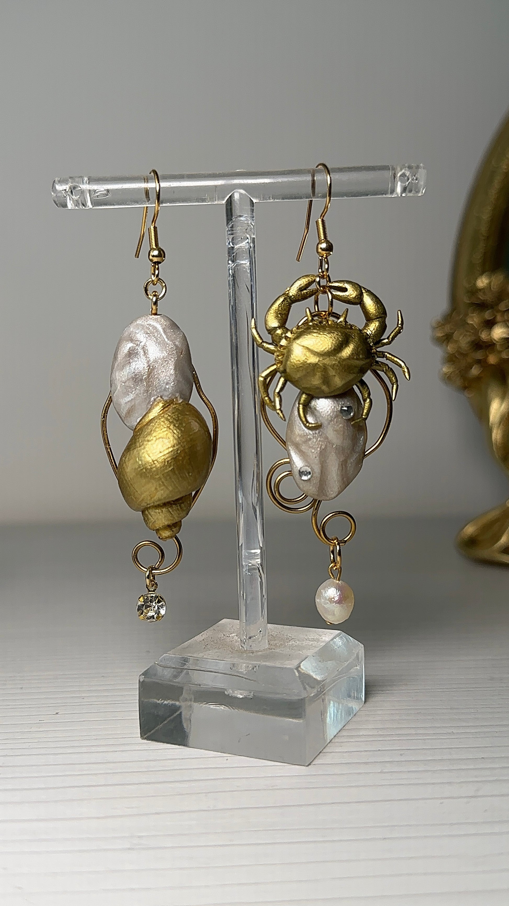 Hand holding a pair of asymmetrical sea-themed earrings featuring a sculpted crab and a golden seashell, with freshwater pearls and gold details.