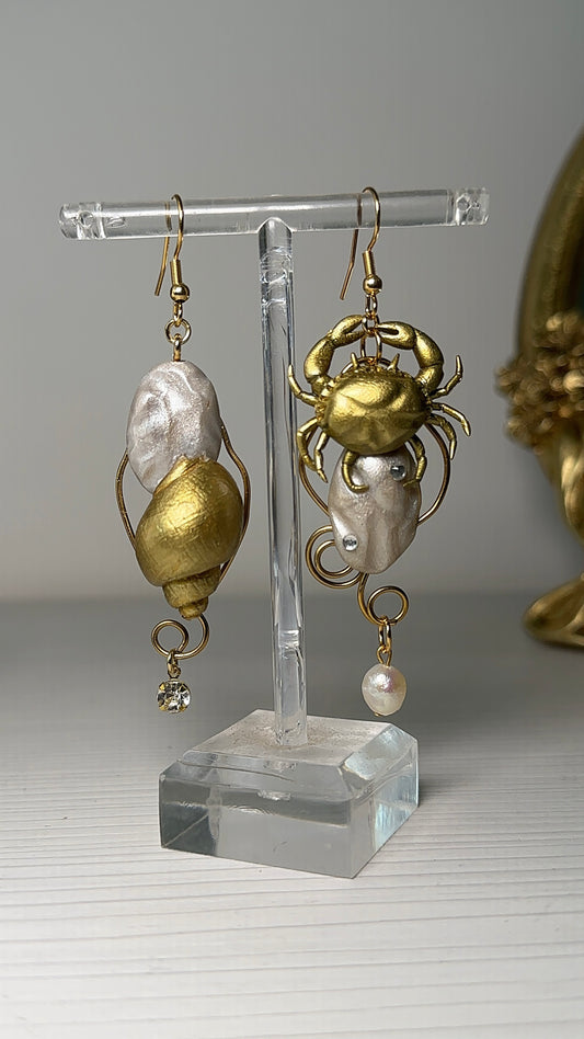 Hand holding a pair of asymmetrical sea-themed earrings featuring a sculpted crab and a golden seashell, with freshwater pearls and gold details.