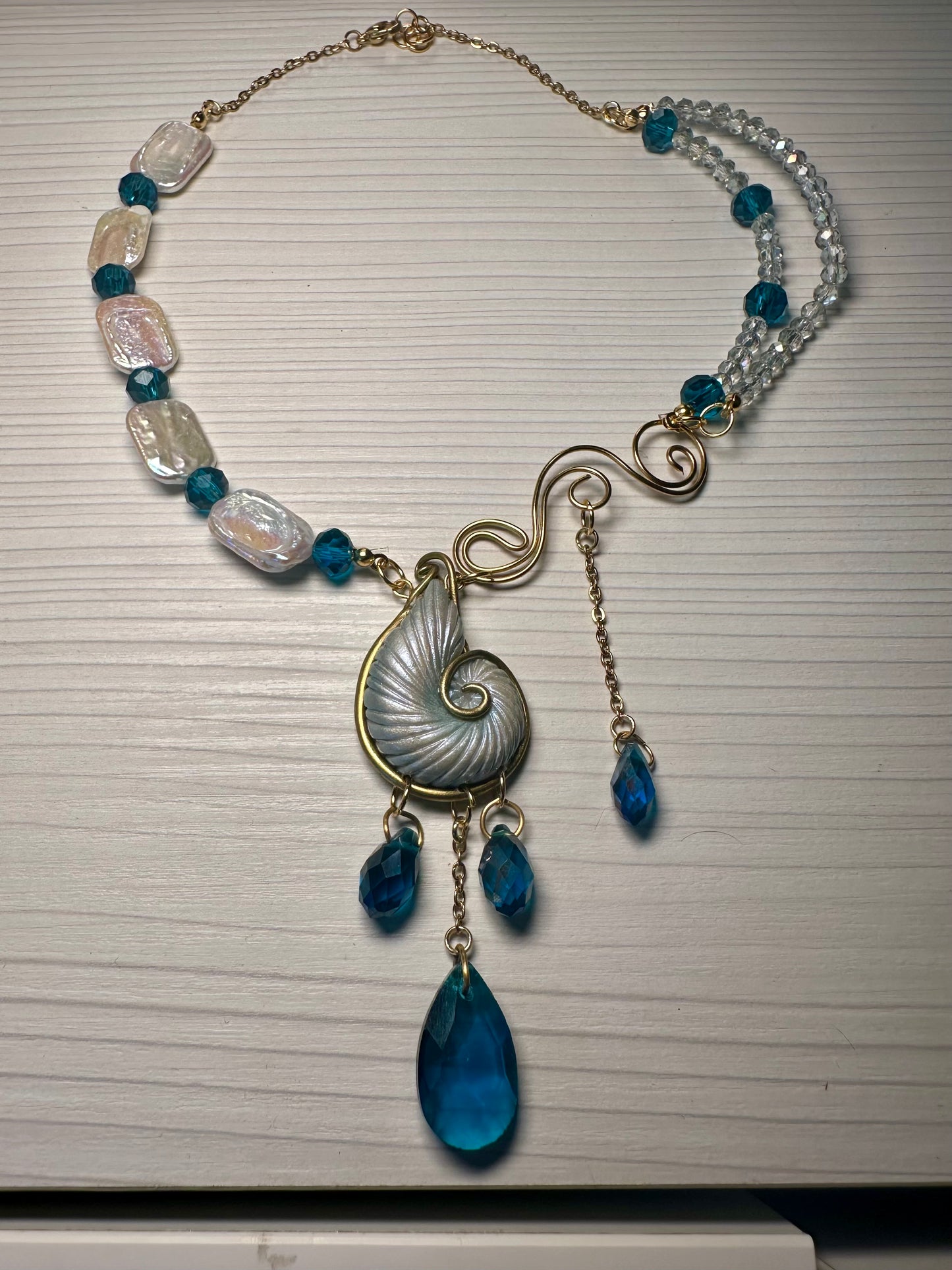 Handcrafted asymmetrical statement necklace featuring nautilus shell, pearls, and teal crystals.