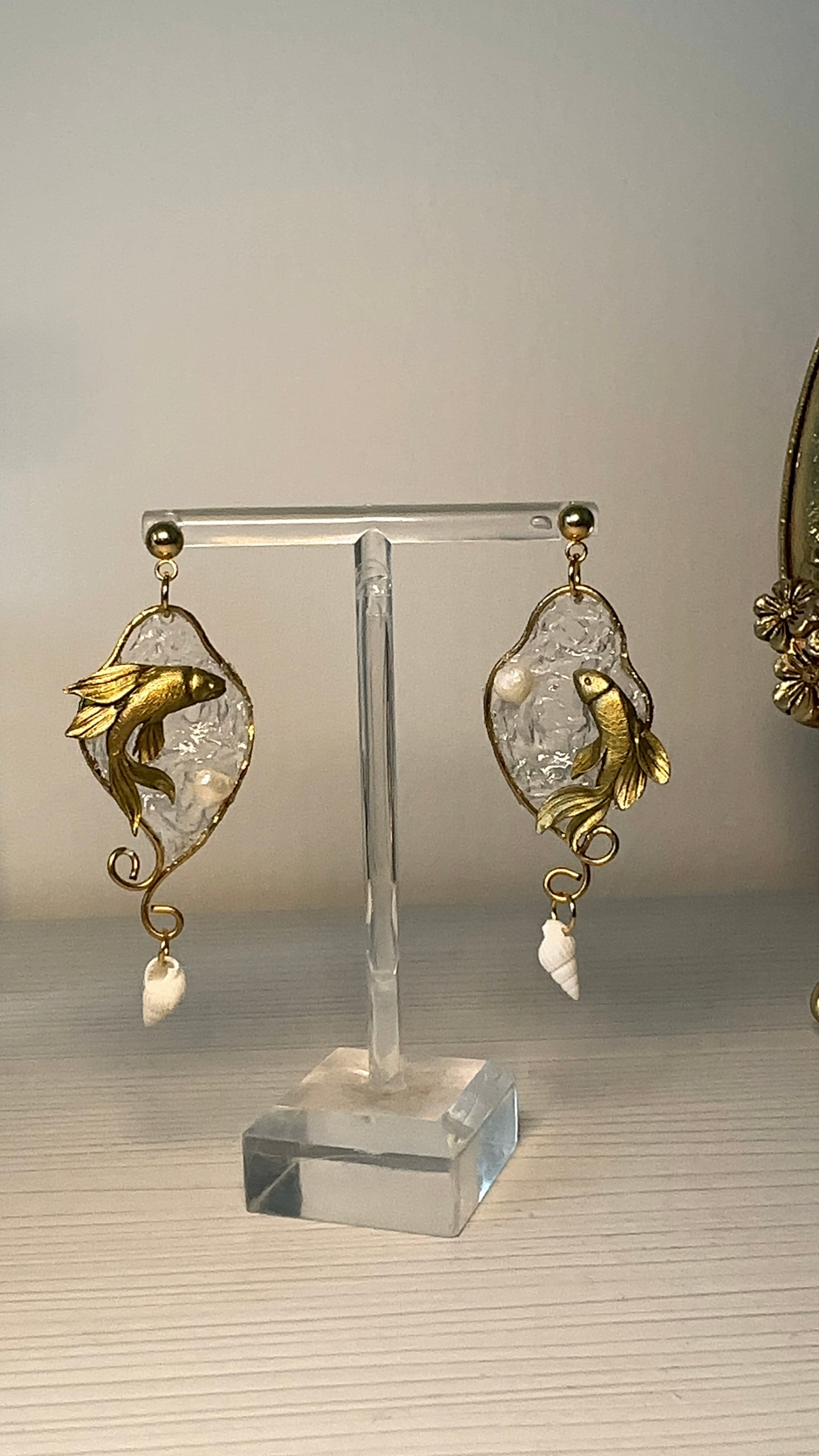Coppia di orecchini artigianali con pesci betta dorati sospesi in una base trasparente effetto mare, decorati con perle e piccole conchiglie pendenti.