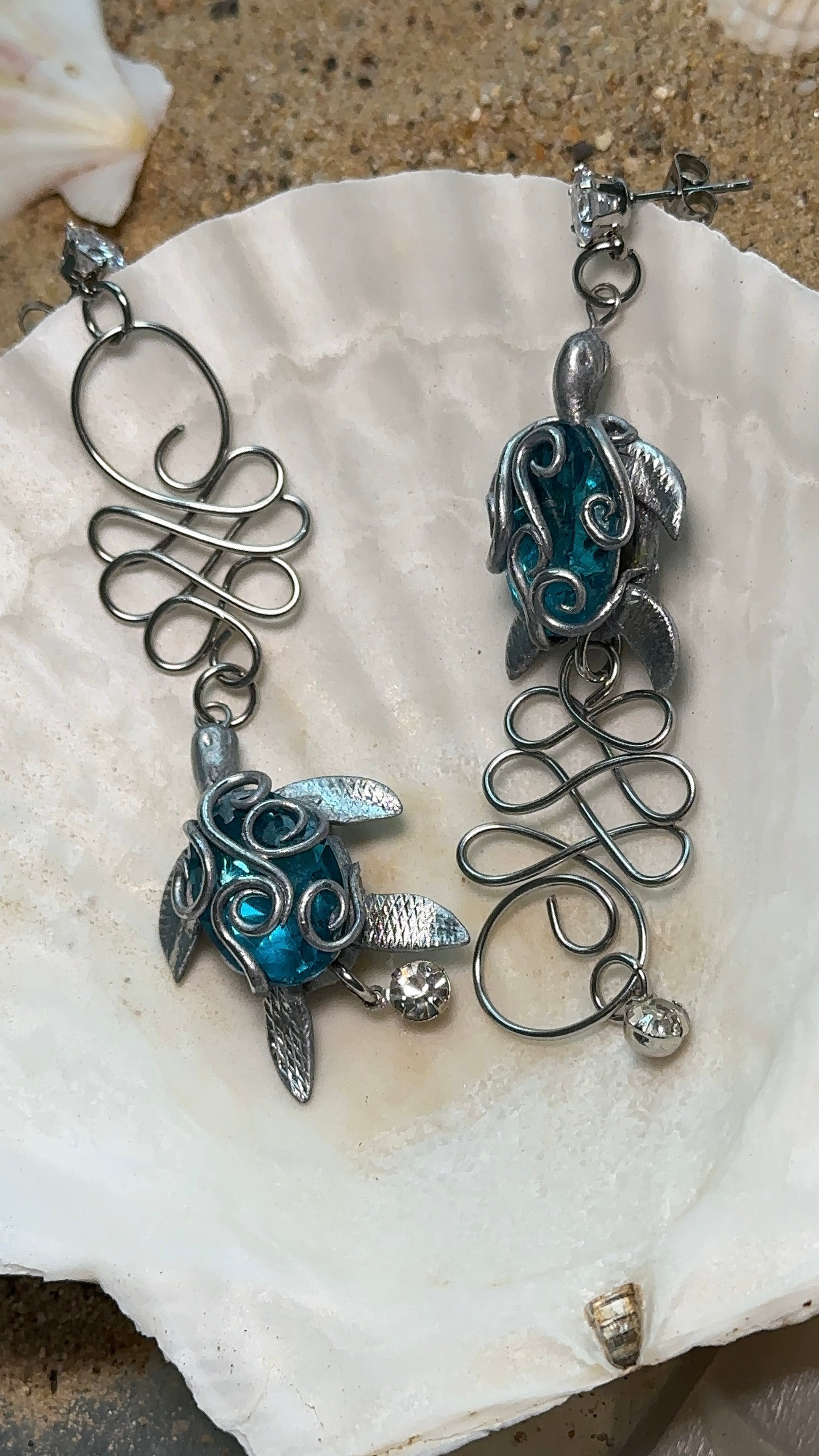 Orecchini artistici con tartarughe blu metallizzate e riccioli scolpiti, fotografati su uno sfondo di conchiglia.