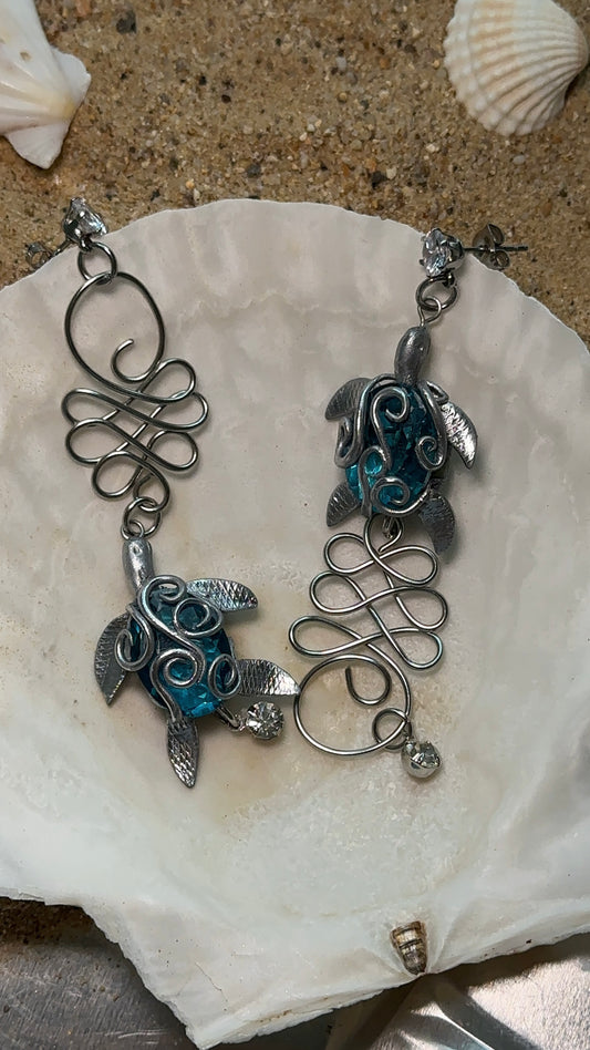 Asymmetrical ocean-inspired earrings featuring hand-sculpted sea turtles in deep blue, wrapped in silver wire filigree.