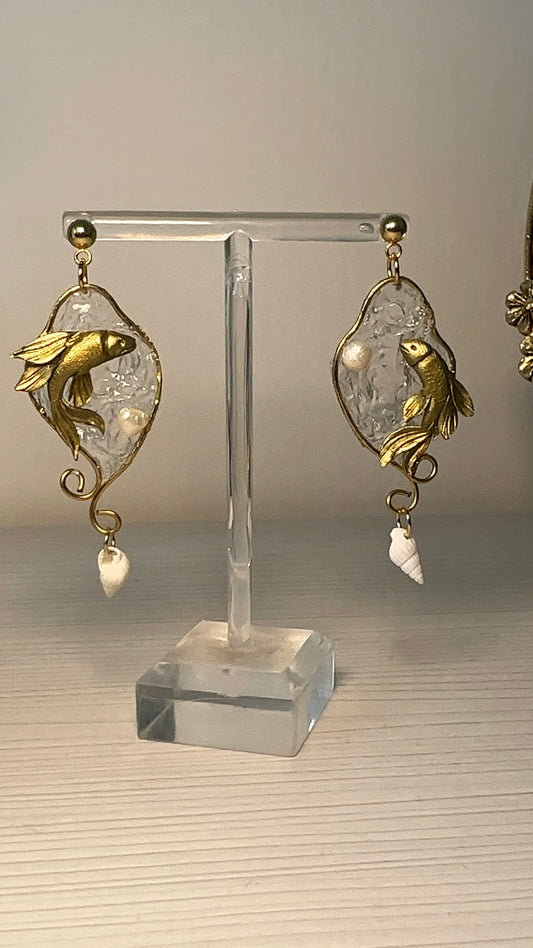 Artistic earrings with sculpted golden fish swimming inside a transparent water-like setting, finished with delicate sea-inspired details.