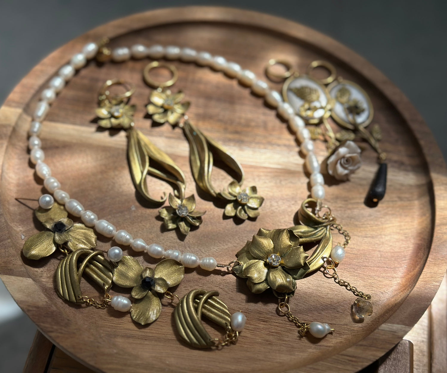 Forever Pieces handmade jewelry collection with pearl necklace, gold sculptural floral earrings, tassel statement earrings and Art Nouveau inspired details on a wooden tray.
