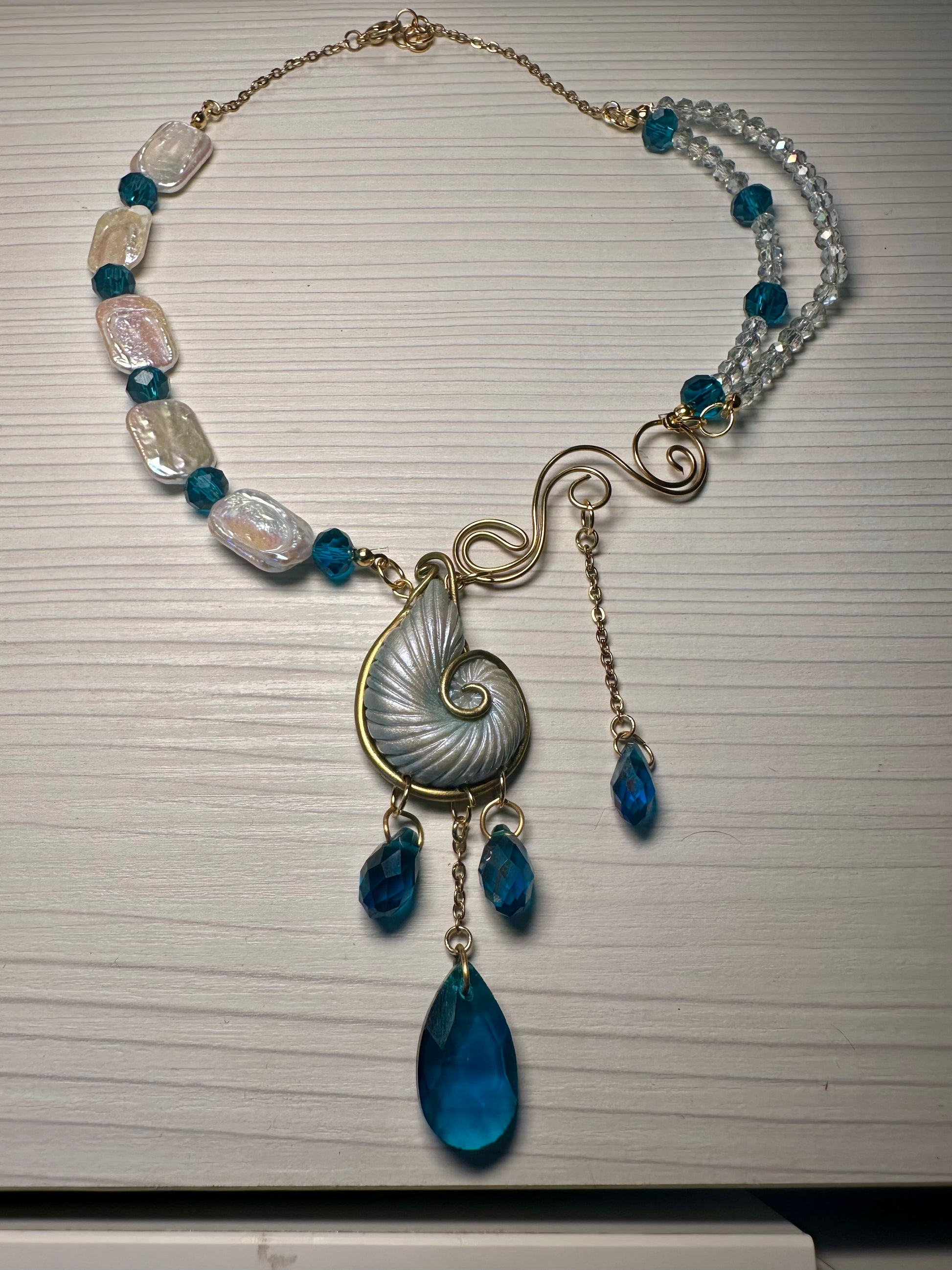 Handcrafted asymmetrical statement necklace featuring nautilus shell, pearls, and teal crystals.