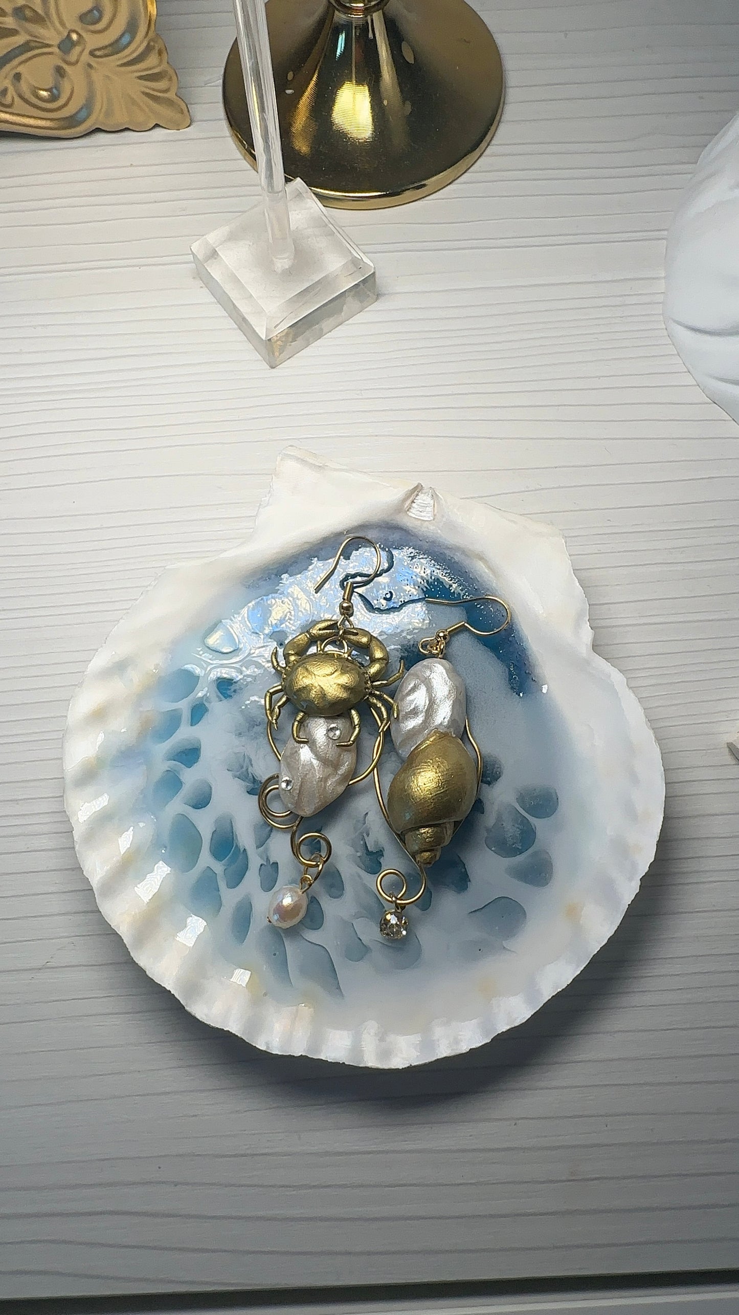 A pair of asymmetrical earrings resting on a handmade resin seashell dish with a water-like effect. The earrings feature a gold crab, pearl-like beads, and delicate marine details — evoking the feeling of treasures sculpted from the ocean floor.
