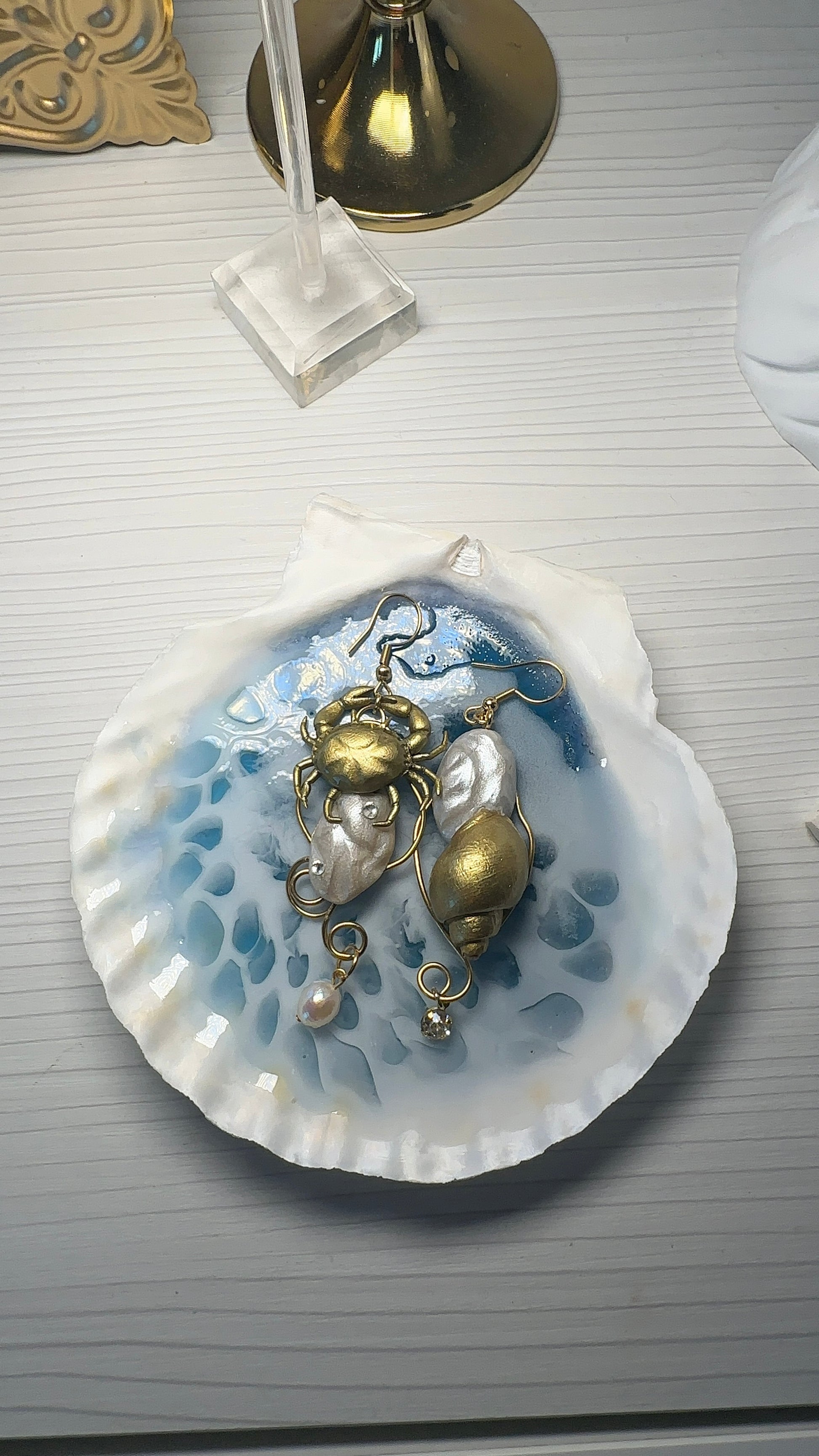 A pair of asymmetrical earrings resting on a handmade resin seashell dish with a water-like effect. The earrings feature a gold crab, pearl-like beads, and delicate marine details — evoking the feeling of treasures sculpted from the ocean floor.