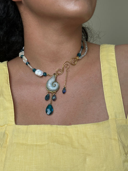     Asymmetrical handmade statement necklace with nautilus shell pendant, blue crystal drops, pearls and gold wire details, styled with a yellow summer dress.